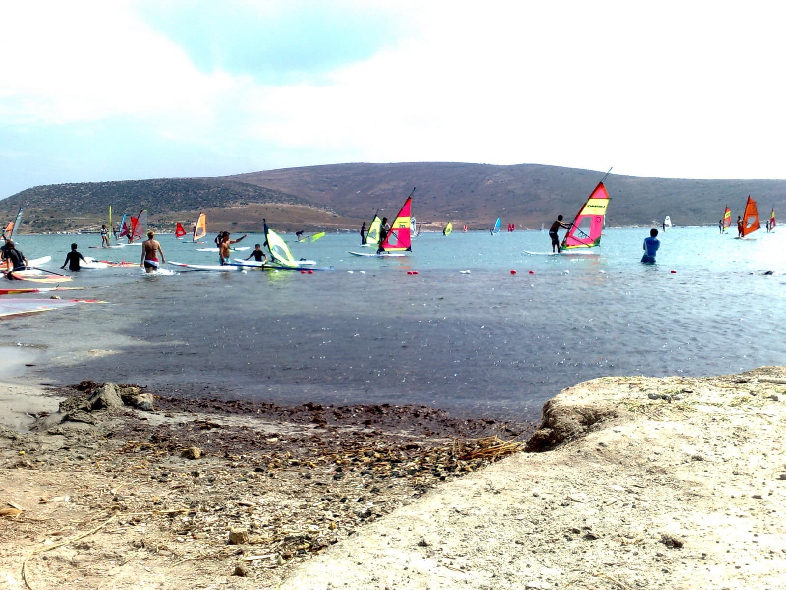 Beginner windsurfers standing in the shallow water of Alacati bay with colorful sails